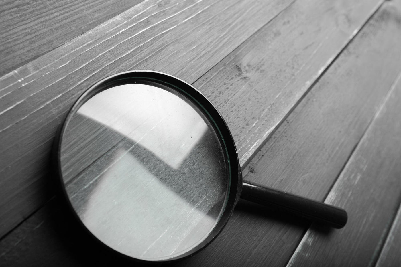 The image shows a magnifying glass placed on a wooden surface, creating a reflection of a window or light source. The black-and-white tone gives it a sleek, investigative, or analytical feel. 