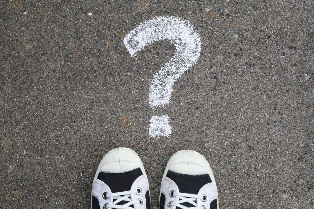 The image features a pair of sneakers standing in front of a large question mark drawn in white chalk on a rough asphalt surface. It conveys a sense of uncertainty, decision-making, or curiosity.