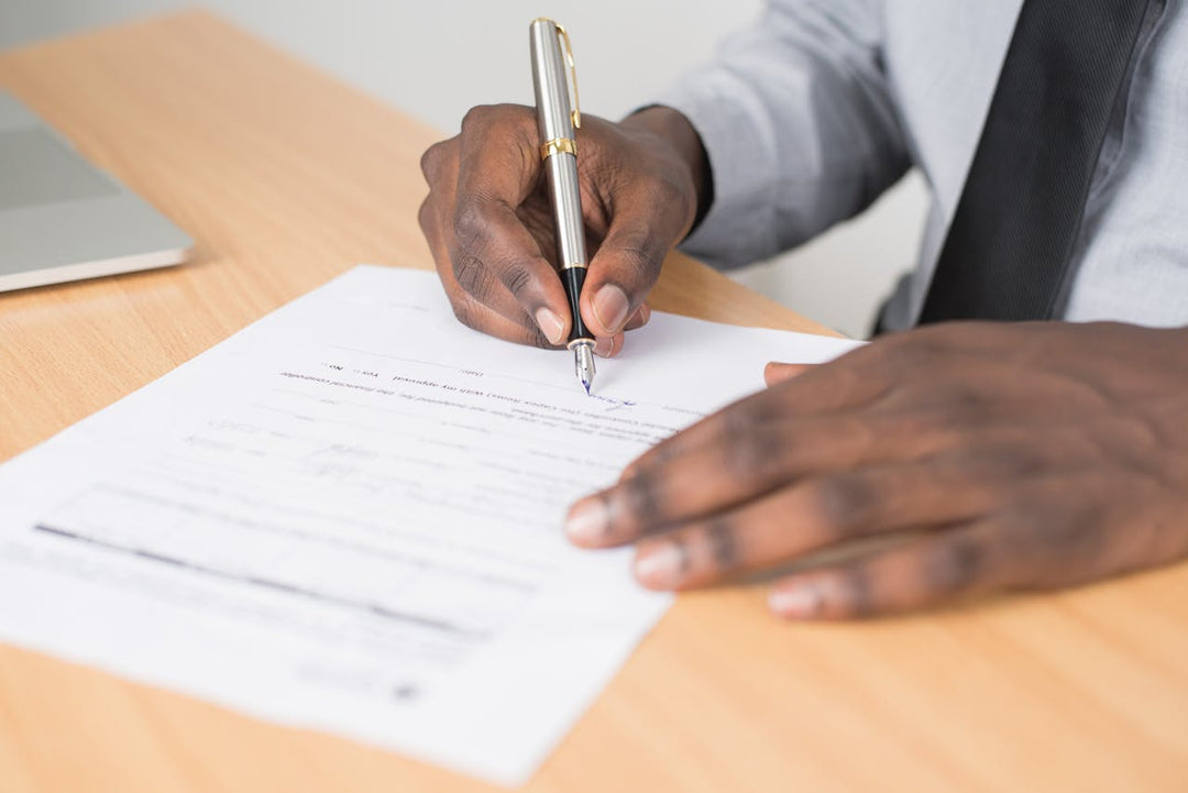 This image shows a person signing a document with a silver and black fountain pen. The individual is wearing a light-colored shirt and a dark tie, and the document is placed on a wooden desk with a laptop partially visible in the background.