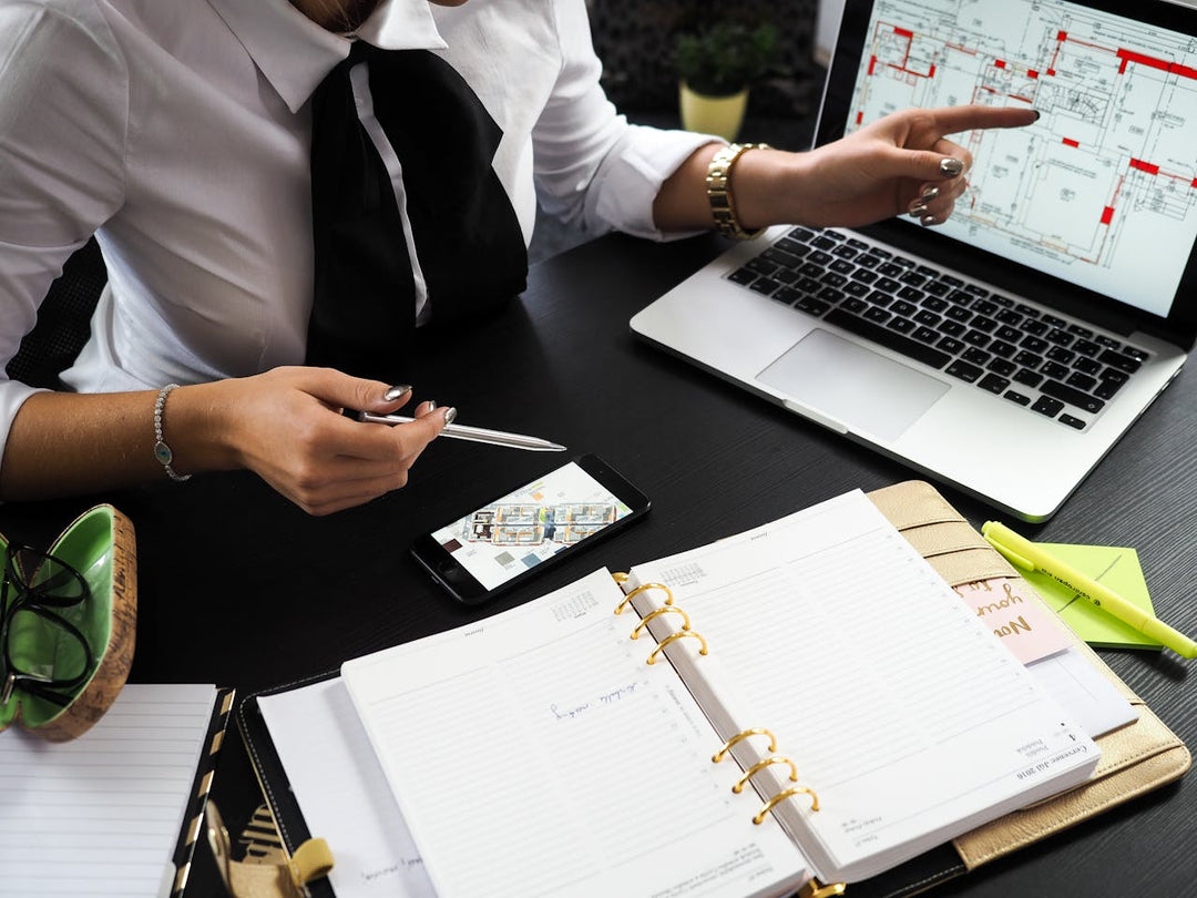 The image shows a professional workspace with a person reviewing architectural plans on a laptop while pointing at the screen. A smartphone displaying a map, an open planner, a pen, and highlighters are also present on the desk.