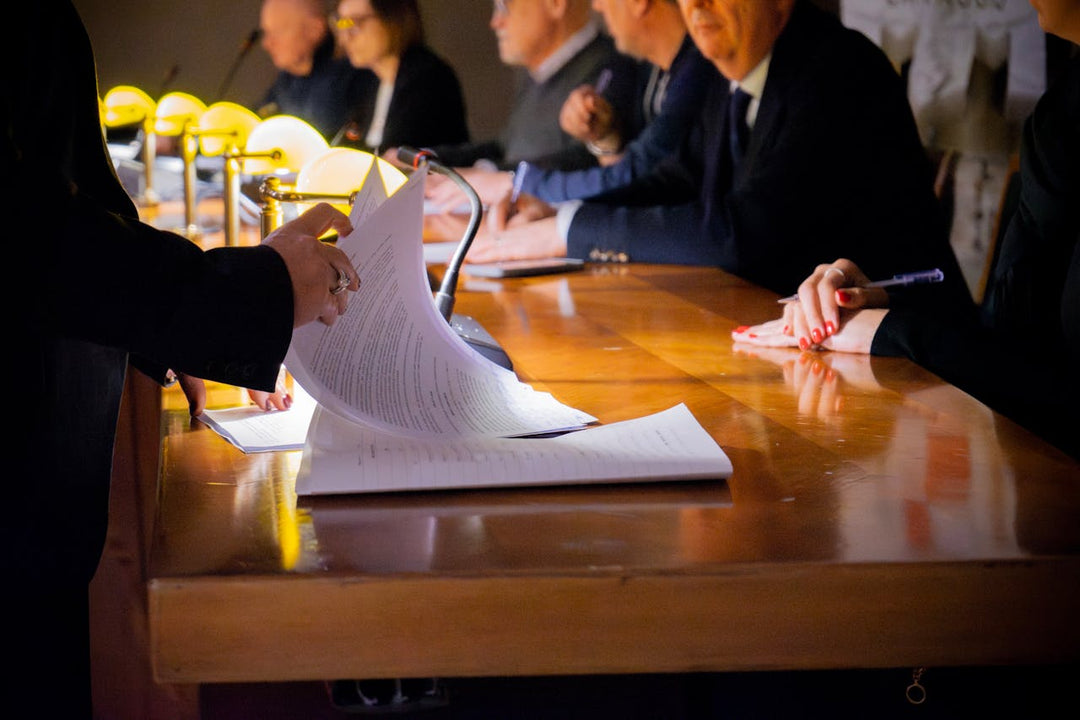 Close-up of a formal meeting with individuals sitting at a wooden table, reviewing and signing documents under warm desk lamps, indicating a professional or legal setting.