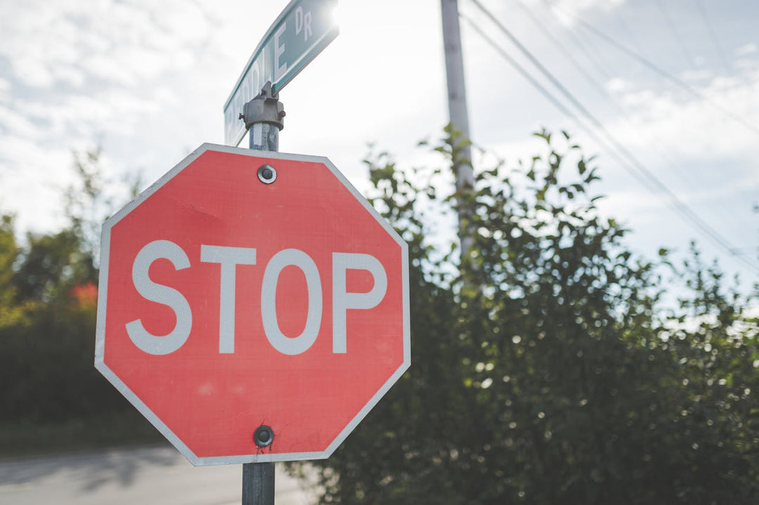 A red octagonal stop sign with bold white lettering stands at an intersection, with a green street sign attached. The background features trees, power lines, and a bright sky with scattered clouds, typical of a suburban or rural area.