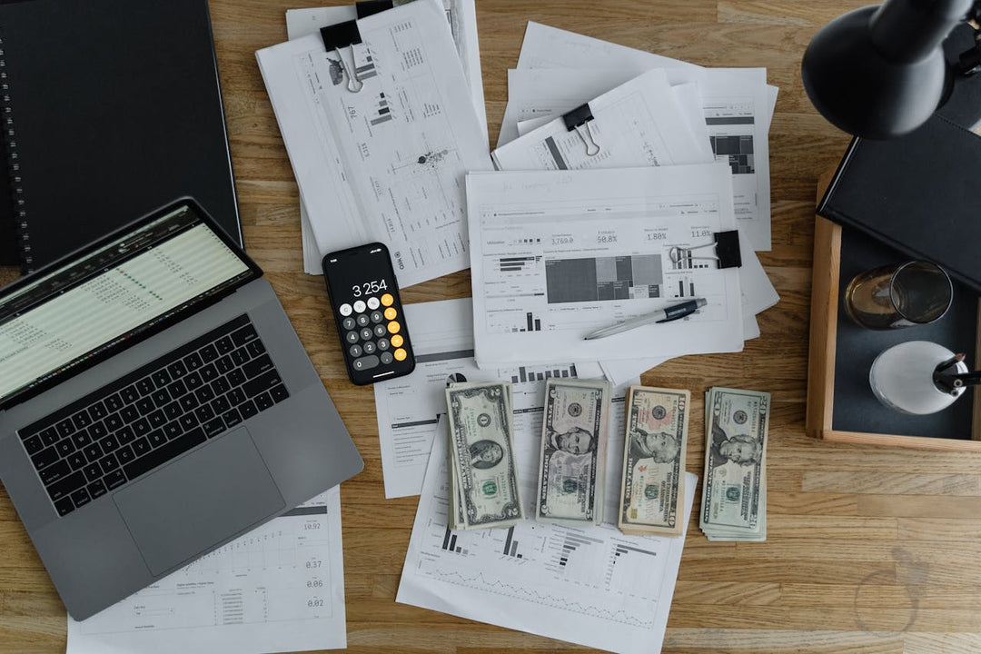 The image shows a desk with financial documents, a MacBook, a phone displaying "3,254" on a calculator app, and stacks of cash.